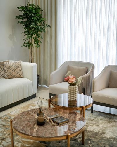 Beige living room with sofa, ribbed armchairs, and nested round brass coffee tables under sheer curtains