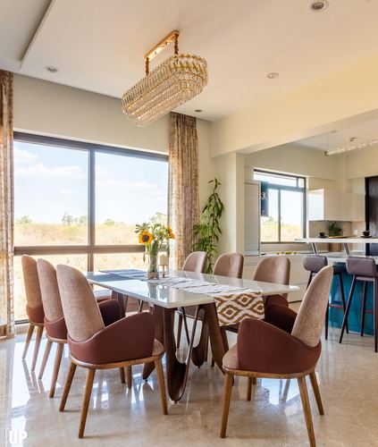 Dining table with mauve chairs under crystal chandelier beside large window, kitchen island beyond