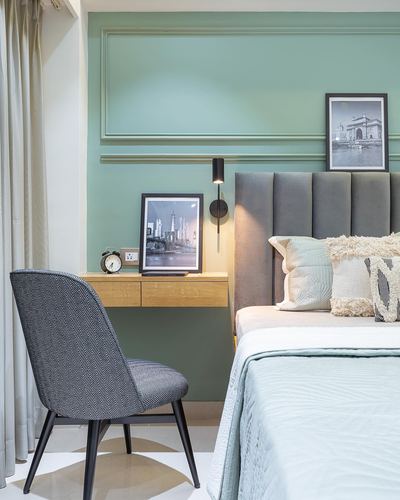 Sage green bedroom wall with molding, floating wood desk, framed prints, and grey channel-tufted headboard