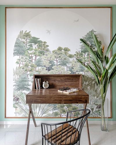 Wooden writing desk with chess set against framed forest mural wallpaper and wire chair