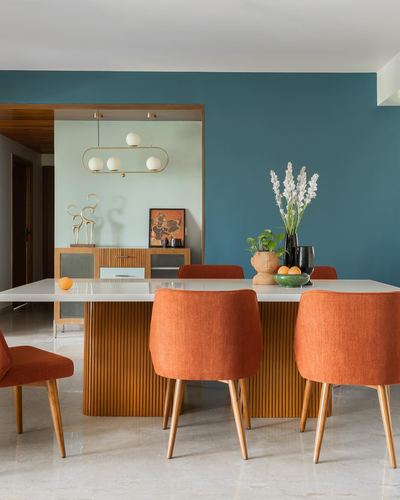 Dining room with orange chairs around marble table against teal wall, fluted wood sideboard beyond