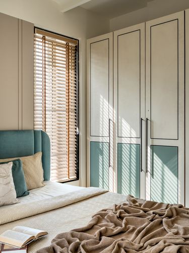 Bedroom with teal upholstered headboard, wooden blinds, and paneled wardrobe in warm sunlight