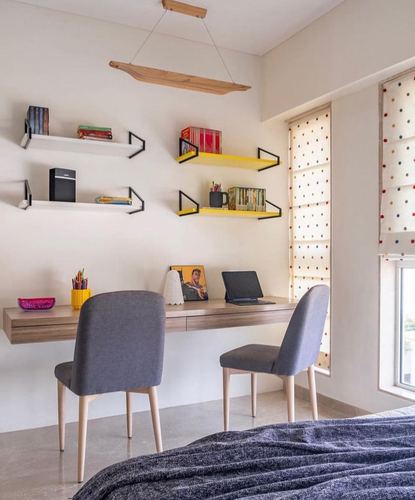 Study nook with floating desk, two grey chairs, and colorful wall shelves under a wooden pendant light