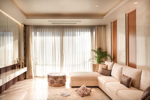 Beige living room with sectional sofa, patterned pouf, and sheer curtained wall glowing with light