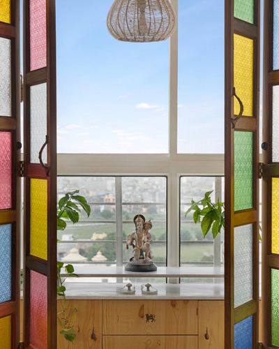 Pooja shelf with deity idol framed by colorful stained glass folding doors and city skyline view
