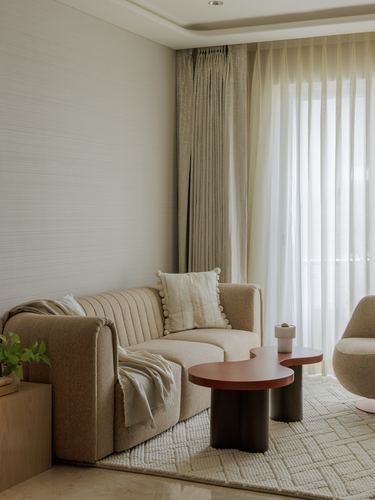 Beige living room corner with channel-tufted sofa, sheer curtains, and sculptural coffee tables