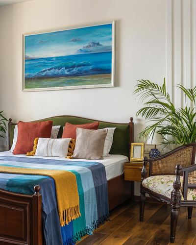 Bedroom with green velvet headboard, striped throw, seascape painting, and cane armchair beside plant