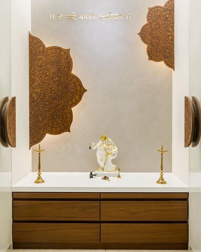 Pooja unit with carved wooden mandala panels, white marble idol of Radha Krishna, and brass diyas on a white countertop above wooden drawers