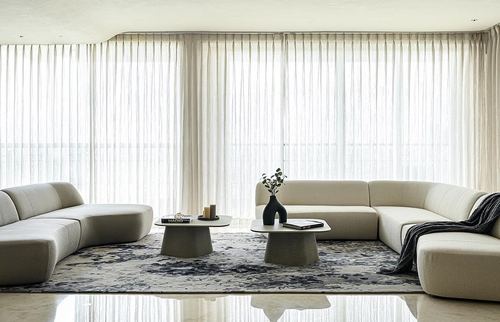 Beige sectional sofas around two round stone coffee tables in a sheer-curtained living room