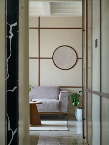 View through a dark stone doorway into a minimalist living room with a mauve armchair and circular mirror