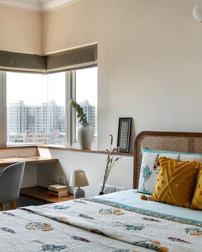 Bedroom corner with cane headboard, floral quilt, and window desk overlooking city skyline