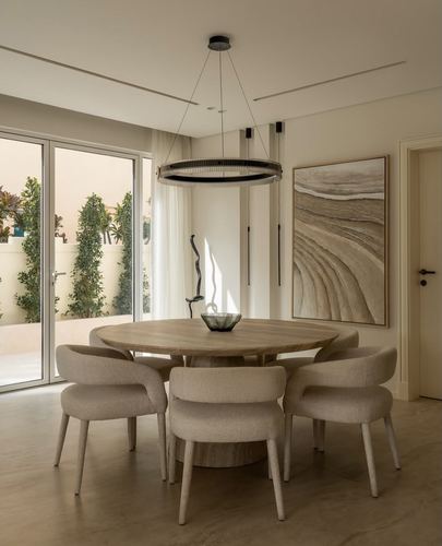 Round wood dining table with beige upholstered chairs under a circular pendant light, glazed doors to garden