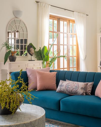 Teal velvet channel-tufted sofa with pink pillows beneath an arched wooden window in a Goan living room