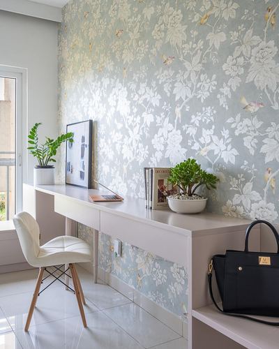 White desk against floral wallpaper wall with chair, plants, and a handbag beside a glass door