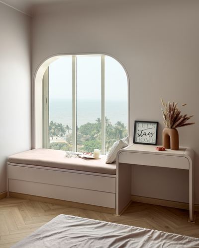 Arched window seat with pink cushion overlooking the sea, beside a small console table with vase and framed print