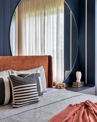 Bedroom corner with rust velvet headboard, striped cushions, and large round mirror against navy fluted wall