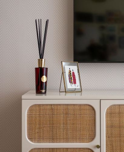 Cane-front cabinet topped with a reed diffuser and small framed art print against herringbone wallpaper.