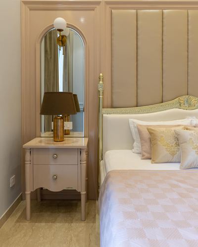 Blush pink bedroom corner with arched mirror, marble-top nightstand, and carved bed with tufted headboard.