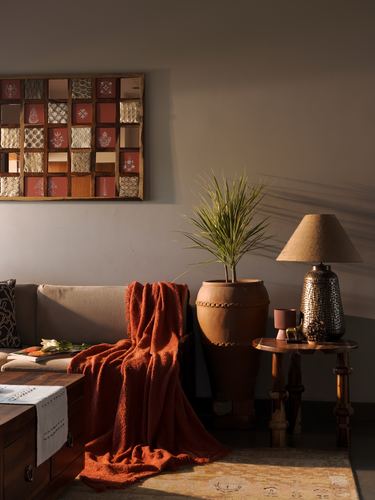 Sunlit living room corner with terracotta pot, patchwork wall art, and orange throw on sofa