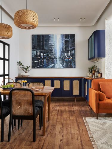 Dining space with cane pendant lights, navy cane sideboard, city painting, and orange armchair beyond