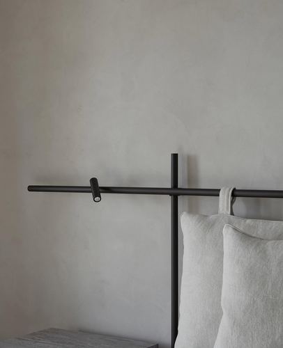 Black metal headboard frame with small reading light against textured plaster wall, linen pillows below