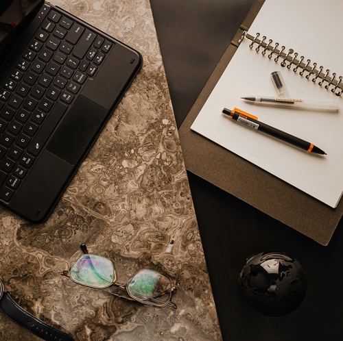 Flatlay of laptop keyboard, notebook and pens on marble and black desk surfaces