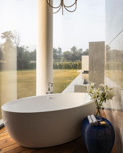 Freestanding oval bathtub beside floor-to-ceiling glass overlooking a lawn, with a blue ceramic side table holding flowers