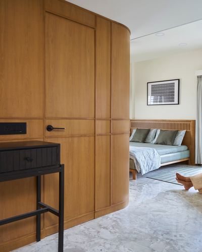 Curved oak wardrobe wall opens into a bedroom with slatted headboard and marble flooring