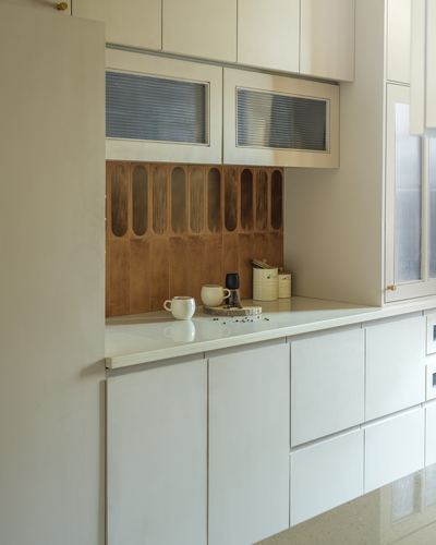 Cream kitchen cabinetry with fluted wood backsplash, reeded glass cabinet fronts, and mugs on a stone counter.