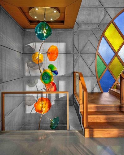 Multicolor glass sculpture chandelier hangs in a concrete stairwell beside a stained glass round window and wooden staircase
