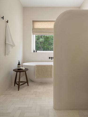 Minimalist beige bathroom with freestanding tub, arched plaster wall, herringbone tile floor, and window to trees