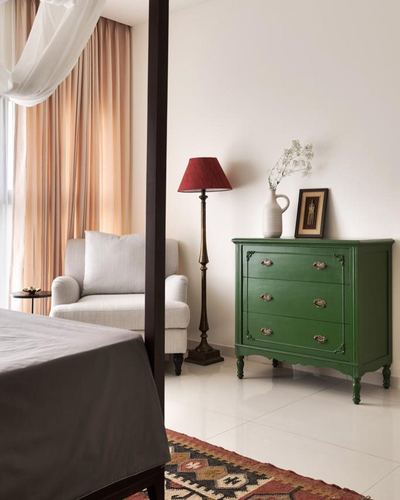 Green painted vintage chest of drawers with brass handles beside a red-shaded floor lamp in a bedroom