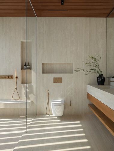 Minimalist stone bathroom with wood-slat ceiling, striped sunlight, wall-hung toilet and brass fittings