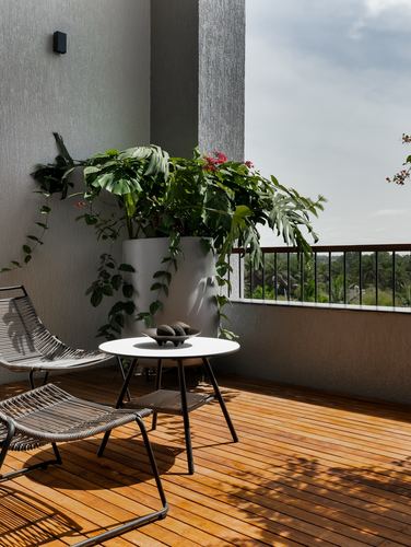Wooden deck balcony with rope lounge chairs, round table, and large potted plant against textured wall.