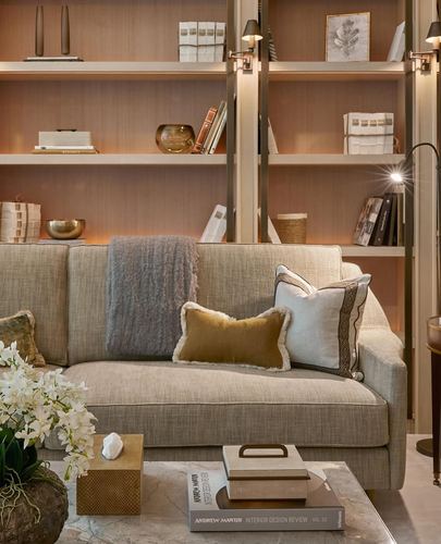 Beige sofa with layered cushions in front of a warm-lit built-in bookshelf and marble coffee table