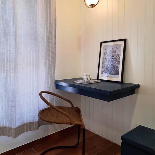 Wall-mounted navy desk with framed art and cane chair by curtained window