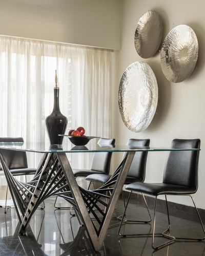 Dining table with sculptural metal base, glass top, black chairs, and textured silver wall discs