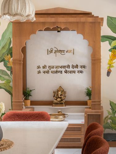 Wooden pooja unit with scalloped arch, Sanskrit text, and brass Ganesha idol, dining chairs in foreground