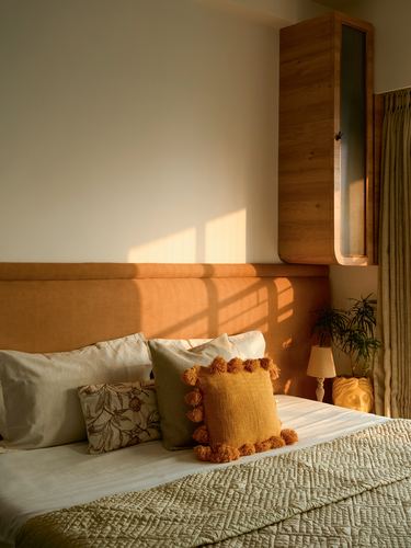 Sunlit bedroom with ochre upholstered headboard, wooden wall cabinet, and tasseled mustard cushion