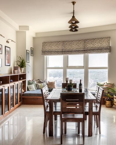 Wooden dining table under a tiered brass pendant light beside a window seat overlooking the city