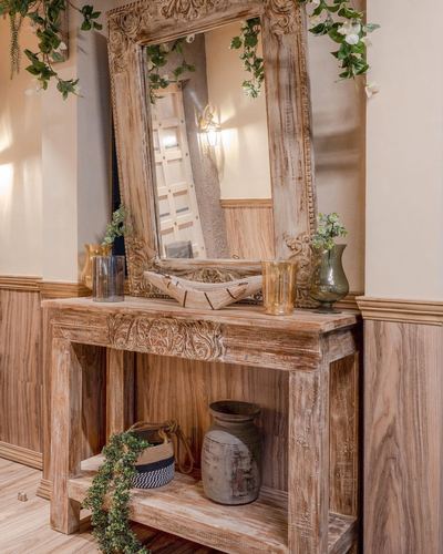 Carved wooden mirror above distressed console table styled with vases and ceramic jug in foyer