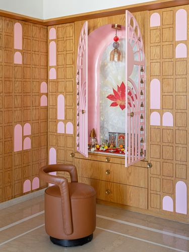Pink arched pooja unit set into wood-panelled wall with brass bells and leather stool