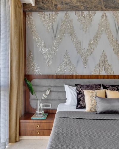 Bedroom corner with tufted grey headboard, wood nightstand, and patterned metallic wall panel