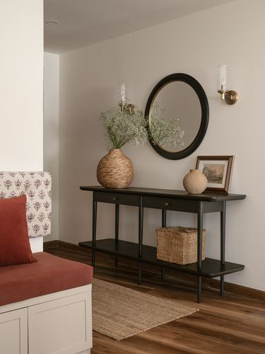 Entryway console table with round mirror, sconces, woven vase, and jute rug beside a patterned bench