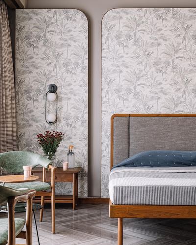 Bedroom with arched botanical wallpaper panels, wood bed, sconce, and green chairs beside a wooden side table.