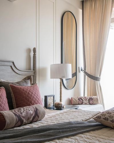 Bedroom corner with carved wooden headboard, pink bolster pillows, oval mirror and sheer curtains