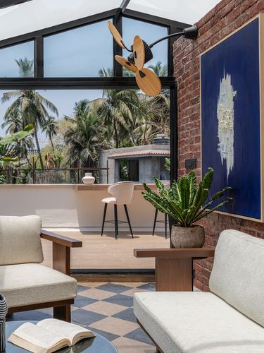 Living room with exposed brick, blue artwork, and glass gable roof opening to a palm-tree balcony