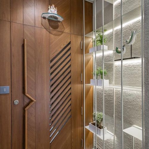 Wooden entrance door with diagonal fluted panel beside a metal shelf screen with plants and textured stone wall