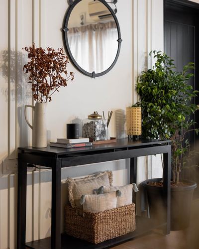 Black console table under round mirror with dried branches, plants, and woven basket in foyer