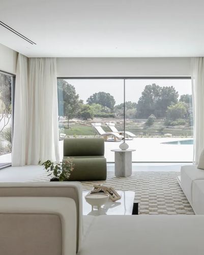 Neutral living room with curved green chair and marble table facing floor-to-ceiling window overlooking pool